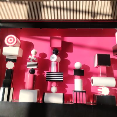 An eye-level indoor photograph captures a modern, abstract art installation or retail display, located within Stockholm-Arlanda, Sweden. The scene is dominated by a vibrant hot pink background wall, against which several multi-component structures are arranged horizontally. These structures consist of stacked geometric shapes, including cylinders, cubes, rectangular prisms, and spheres, rendered in a palette of white, black, and various shades of pink. Specific shapes feature patterns such as concentric pink and white circles, or alternating black and white, or pink and black horizontal stripes. Strong directional lighting from the upper left casts prominent, crisp shadows of the objects onto the pink wall. Above the main display, a horizontal black band is visible, with several small, recessed circular lights embedded within it. Higher in the frame, a ceiling structure composed of numerous light-colored wooden slats, arranged vertically, extends across the top. The entire display rests upon a dark, flat base surface that occupies the bottom portion of the image. No individuals are present in the scene.