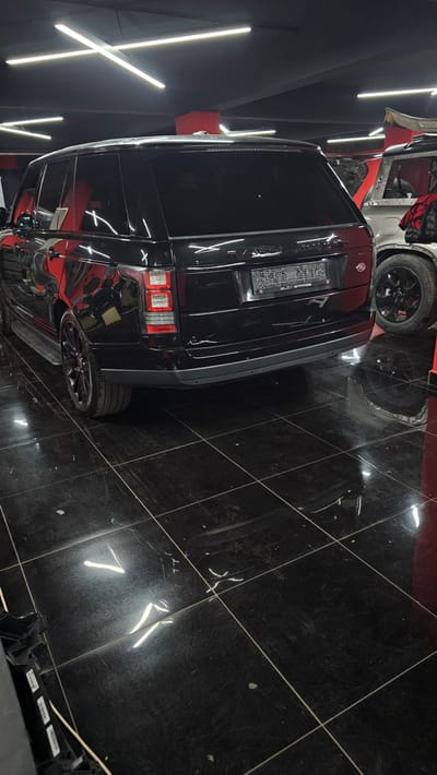 A black Range Rover is parked in a dimly lit garage with a shiny black tiled floor. The vehicle is facing away from the camera, with its rear visible. The garage ceiling is black and features several bright, linear fluorescent lights arranged in an 'X' pattern. Another vehicle, possibly a silver SUV, is partially visible to the right of the Range Rover, with its wheel and fender showing. The scene is clean and organized, suggesting it might be a detailing shop or a private garage. There is no indication of any people or activity in the image. The overall impression is one of automotive care or storage.
