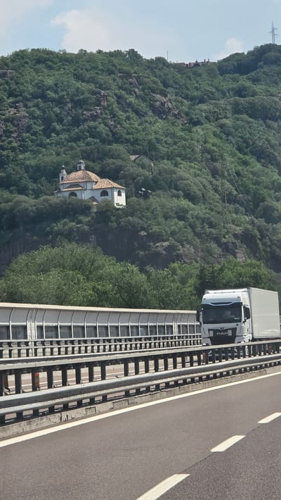 The image captures a white semi-truck driving on a highway with a lush green mountainside in the background. On the mountainside, a white building with a terracotta-tiled roof, possibly a church or chapel, is visible. The highway features metal guardrails and a sound barrier along the side. The sky is partly cloudy. The scene appears to be a scenic route, likely near Bolzano, Italy, given the characteristic mountainous terrain and architectural style.