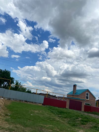 The image depicts a rural scene under a cloudy sky in Taganrogsky, Russia. The foreground is dominated by a lush green grassy field, with some dry vegetation and patchy soil. A gray metal fence runs horizontally across the field, with a dark red metal fence behind it, partially obscured by trees. A house with a brick exterior and a teal roof is visible beyond the red fence, featuring a chimney and a window. Yellow pipes, likely gas lines, are prominent throughout the scene, running along the fences and the side of the house. Overhead, a network of black power lines stretches across the frame, crisscrossing against a backdrop of dramatic clouds.
The sky is a mixture of bright blue patches and large, heavy gray and white clouds, suggesting a day with changing weather, possibly a mix of sun and approaching rain. The lighting indicates it is daytime, likely late morning or early afternoon. The scene has a quiet, everyday feel, with no people or animals visible. The presence of gas lines and the style of the house and fences point to a residential area in a suburban or rural Russian setting. There is no visible text in the image.