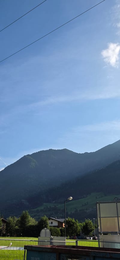 The image depicts a mountainous landscape under a clear blue sky with thin, wispy clouds. Two power lines traverse the upper portion of the frame. In the mid-ground, a dark, forested mountain range dominates the scene, with patches of lighter green suggesting open fields or lighter foliage. At the base of the mountains, nestled among trees, are several residential houses. A street lamp stands to the right of center, its light diffused by the bright sky. In the foreground, a green grassy field is visible, bordered by a metal fence. A portion of a large, open-top refuse container is prominent in the bottom right corner of the frame. The scene appears to be located in or near Anger, Austria, given the characteristic Alpine terrain and architecture. There are no people or overt actions visible in the image.