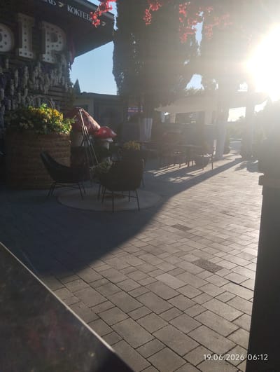 The image captures a sunlit outdoor seating area, likely part of a restaurant or cafe, in Kiparisnoe, Ukraine. The foreground shows a paved patio with a distinct shadow cast by overhead structures or trees, indicating a sunny day. Two dark, modern-style chairs are visible on the left, situated on a circular rug. Behind these chairs, a large, woven basket overflows with yellow flowers, and two decorative red and white mushroom sculptures are placed nearby. In the background, several tables and chairs are set up, some under a covered structure. The bright sunlight creates a lens flare effect in the upper right quadrant of the image. The overall scene suggests a tranquil and inviting outdoor space.
