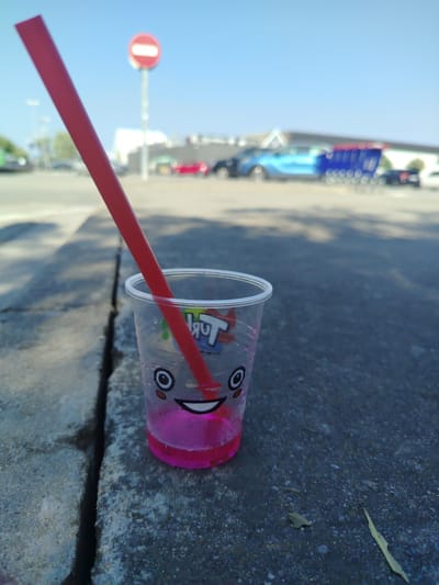 A disposable plastic cup containing pink liquid and a red straw rests on the edge of a cracked concrete curb. The cup has a cartoon face printed on it. In the background, a street, cars, shopping carts, and a "no entry" traffic sign are visible. The scene is set in a city, possibly Santa Susanna, Spain, under a clear blue sky.