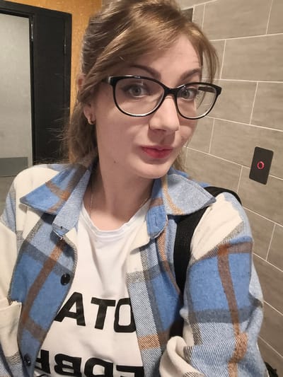 A young woman with auburn hair and glasses is taking a selfie in what appears to be an elevator. She is wearing a white t-shirt with black lettering that reads "ARATORI" and a blue and white plaid shirt over it. Her hair is pulled back loosely, and she is wearing small hoop earrings. Behind her, a grey tiled wall is visible, with a black elevator call button that has a red ring around the center. The overall environment suggests an indoor, possibly public, space.