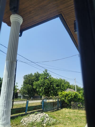 The image shows a view of a rural property on a clear day. In the foreground, a large white fluted column, partially wrapped in plastic, stands to the left. To the right, the edge of a dark frame obstructs the view. A wooden soffit extends overhead.
Beyond the column and frame, a metal gate with blue uprights and green panels, decorated with ornate scrollwork, spans across the middle ground. Behind the gate, a grassy yard with scattered debris is visible. Further back, a dense line of green trees and foliage obscures a house with a dark door and windows. Power lines are strung across the sky. To the right of the gate, a rustic wooden fence encloses an area with lush green vines. The overall scene depicts a residential area in the village of Grinăuți, Moldova.
