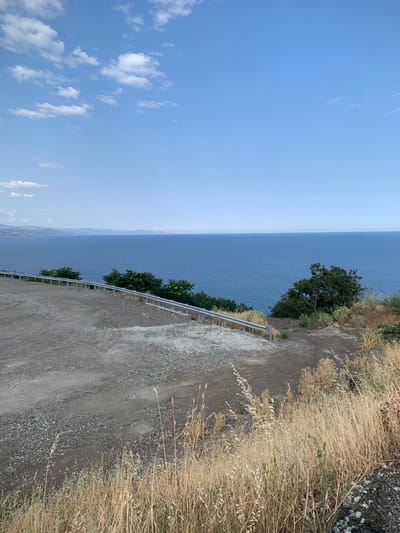The image depicts a scenic overlook of the Black Sea, likely near Lazurnoe, Ukraine. In the foreground, a gravelly, unpaved area slopes downwards, bordered by a metal guardrail. Dry, tall grasses are prevalent on the right side of the frame. Beyond the guardrail, lush green trees and bushes dot the landscape. The expansive, deep blue Black Sea stretches to the horizon, meeting a clear, light blue sky dotted with a few wispy white clouds. In the distance, a hazy, mountainous coastline is visible. There are no people or animals in the frame. The scene is tranquil and natural.