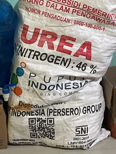The image shows a large white woven bag of UREA (NITROGEN) fertilizer with red lettering. The bag has various markings and text printed on it, including "PU UP INDONESIA HOLDING COMP" and "INDONESIA (PERSERO) GROUP". There is also a QR code and text that appears to be a date and location: "DESEMBER 2025" and "PALEMBANG-INDONESIA". The fertilizer bag is positioned in what appears to be a storage area, with glimpses of other objects like cardboard boxes and a blue plastic container in the background. A small blue and white spigot is attached to the side of the bag, suggesting it might be repurposed. The scene is lit, and the focus is on the bag and its text. There are no people or animals visible in the image. The location is identified in the metadata as Tambakreja, Indonesia.