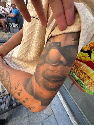 The image shows a close-up view of a person's arm covered in tattoos, with a portion of their torso and clothing visible. The tattoo on the arm appears to be a realistic portrait of a face with sunglasses. In the background, out of focus, are two men sitting at a table outside, with one wearing a blue t-shirt and sunglasses. The setting appears to be an outdoor cafe or restaurant, with tables and chairs visible. The overall impression is casual and relaxed. This photograph was taken in the city of Deventer, Netherlands.
