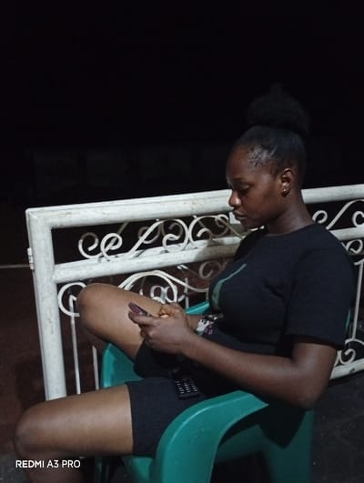 A young woman is sitting on a green plastic chair in an outdoor setting. She's engrossed in her phone, which she holds in her hands. She wears a dark t-shirt and shorts. Her hair is styled in a bun. A white, ornate metal railing serves as a backdrop behind her. The lighting is dim, suggesting it might be evening or night. There is a remote control on her lap.