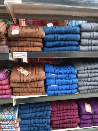 Neatly stacked towels fill store shelves. Various colors are displayed, including brown, blue, gray, pink, burgundy, and purple. Some towels have tags attached. The towels appear to be made of a soft, textured material with visible ridges. A price tag of 29,900 is visible on the middle shelf. The shelves are grey.