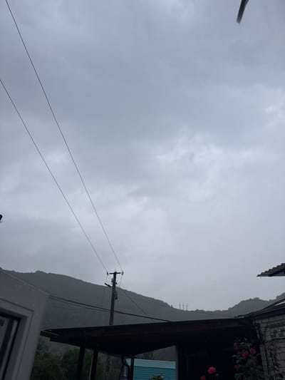 The image depicts an overcast sky with heavy clouds, suggesting rainy weather. In the foreground, a portion of a white window frame is visible on the left. Power lines stretch diagonally across the frame, connecting to a utility pole with visible insulators. Beyond the power lines, a dark, covered structure with a visible roof is present. Further in the background, a lush green, tree-covered mountain dominates the landscape, its slopes shrouded in mist. Electrical towers are faintly visible on the ridge of the mountain. The overall impression is a view from a residential area in Каменномостский, Russia, during a period of rain. There are no people or animals visible in the scene.