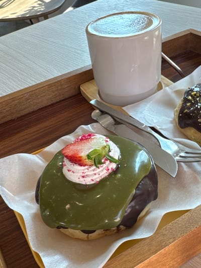 The image depicts a close-up view of a dessert and beverage serving on a light-colored table in San Phra Net, Thailand. No individuals are visible. A wooden serving tray occupies the central and lower portions of the frame. On the tray, a white ceramic mug with a light brown rim containing a frothy beverage (likely coffee or latte) rests on a small, light-colored wooden coaster. Adjacent to the mug, a silver knife and fork are positioned on white parchment paper. Two donuts are presented on the tray. The prominent donut in the foreground features a dark chocolate glazed base, a thick green (possibly matcha) glaze topping, a dollop of pink-tinted whipped cream, a half-strawberry slice, and pink sprinkles. A second donut, partially visible to the right, is covered in dark chocolate glaze and garnished with chopped nuts (appearing to be pistachios) and white sprinkles. Both donuts rest on white parchment paper within yellow plastic or ceramic dishes. The table surface is light-toned and textured, with natural light casting shadows in the upper left. The scene suggests a cafe or restaurant setting.