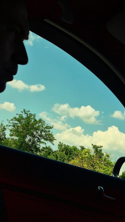 The image captures a perspective looking up through a car sunroof towards a bright blue sky dotted with fluffy white clouds. Lush green trees line the right side of the frame, suggesting an outdoor setting in a natural environment. In the lower left corner, a silhouette of a person's face is visible, looking upwards. The person's head is angled, with their profile in shadow against the bright sky. The car's interior is hinted at by the dark, curved edges of the sunroof and the reddish-brown upholstery on the right side. The overall scene evokes a sense of calm and contemplation, perhaps a leisurely drive or a moment of reflection. The sunlight suggests it is daytime, likely late morning or afternoon, with pleasant weather. The location context of Mihailovca, Moldova, implies a rural or semi-rural setting with abundant greenery. There is no visible text in the image.