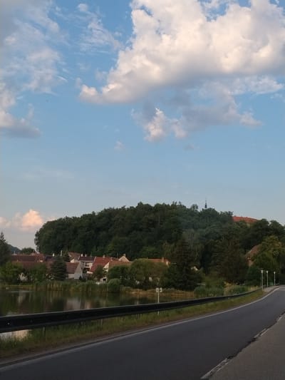 The image depicts a scenic view of the outskirts of Strakonice, Czechia. In the foreground, a paved road curves to the right, bordered by a black metal guardrail and vegetation. Beyond the guardrail, a calm body of water reflects the sky and the trees. On the far side of the water, a small village with red-roofed houses is nestled at the base of a densely wooded hill. A church steeple with a cross is visible rising above the trees on the hilltop. The sky is a clear blue, punctuated by fluffy white clouds. The overall impression is one of a peaceful rural landscape.