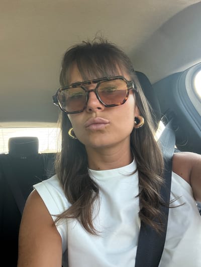 The image captures a woman taking a selfie or being photographed inside a car, set in Le Muy, France. She is seen from the chest up, positioned within the car's interior. She has long, straight brown hair styled with bangs, partially covering her forehead. She wears large, rectangular eyeglasses with a tortoiseshell-patterned frame and golden hoop earrings. Her lips are slightly pursed in a posed expression.

She is dressed in a plain white t-shirt, and a black car seatbelt is visible diagonally across her chest and shoulder. The background reveals elements of the car's interior, including a light-colored headliner or door panel on the left and a darker car seat or headrest on the right. Bright daylight streams through a car window in the upper background, indicating it is daytime. There is no visible text in the image.