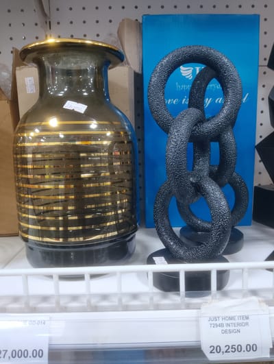 The image displays a retail shelf in what appears to be a home decor store. The foreground features two distinct decorative items. On the left is a large, dark glass vase with gold horizontal stripes. A price tag with the handwritten amount "27,000.00" and some obscured text is attached to the vase. To the right of the vase is a sculpture made of interconnected black rings, resting on a black circular base. Behind the sculpture, a bright blue rectangular panel is visible, with some white text that is partially obscured. The sculpture also has a small white label attached to its base. In the background, cardboard boxes and more retail shelving are visible. Below the shelf, another price tag is visible, indicating "JUST HOME ITEM 7294B INTERIOR DESIGN" and a price of "20,250.00". The lighting suggests an indoor setting, likely during the day. The overall scene is a static display of home goods.