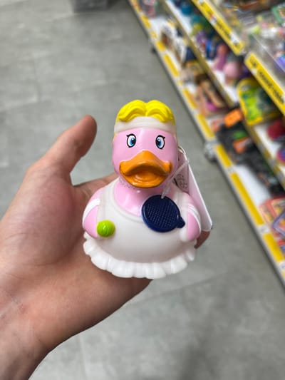 A person's hand holds a pink rubber duck with yellow hair, blue eyes, and an orange beak. The duck has a white ruffled edge around its body, a blue circular detail on its chest, and a small green bulb on its side. The background shows shelves in a store with various colorful items. The floor appears to be tiled. The lighting suggests an indoor setting, possibly a supermarket or toy store, during the daytime.
