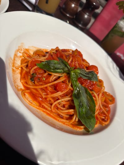 A plate of spaghetti with tomato sauce and basil sits on a white plate. The dish appears to be served outdoors, potentially in a restaurant, given the presence of a salt and pepper grinder and a drink in the background. The location is Guildford, United Kingdom. The plate is positioned at an angle. The sauce includes halved cherry tomatoes. Three basil leaves garnish the dish.