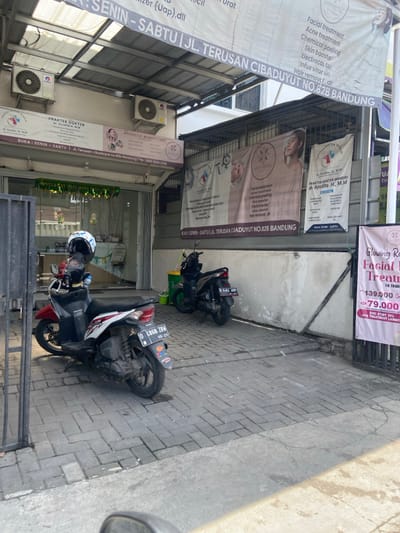 The image shows a storefront in Kecamatan Dayeuhkolot, Indonesia, located at latitude: -6.967, longitude: 107.592. The storefront has a sign that reads "Praktek Dokter dr. Ayudita, H, M,M" and "Aesthetic Beauty Care." Two motorcycles are parked in front of the storefront. There are multiple signs advertising beauty treatments and services, with prices listed. The storefront appears to be a beauty clinic or spa. The environment is an urban area, with paved sidewalks. The motorcycles are parked in the shade of a canopy, and the storefront has a metal gate. The signs are in Indonesian and English. The image is taken from a slightly elevated perspective.