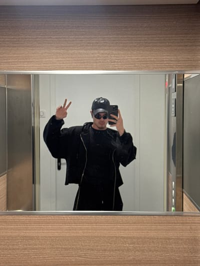 An individual is depicted in a mirror selfie taken inside an elevator located in Asnières-sur-Seine, France. The subject, a male, is dressed entirely in black, consisting of a baseball cap with a metallic or reflective band, dark sunglasses, a zippered jacket, and a t-shirt. He holds a dark-colored smartphone in his right hand, angled towards the mirror to capture the image. His left hand is raised, forming a V-sign (peace sign). The elevator interior is characterized by light wood-grain patterned panels framing the top and bottom of the large, reflective mirror, with a narrow silver trim strip separating the mirror from the panels. A white wall is visible in the reflection behind the subject, along with a silver elevator control panel featuring several illuminated buttons on the right side of the reflected scene.