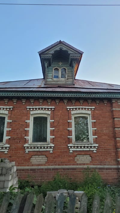 The image captures a low-angle, eye-level view of the upper facade of a two-story brick building in Afoninо, Russia. The building features a steeply pitched, dark-colored roof, likely made of metal, showing signs of rust and wear. Prominently, a dormer window structure with intricate, carved wooden detailing in a pale blue-green hue protrudes from the roof. This dormer has a pointed, decorative roofline with a complex pattern of cutouts. Below the dormer, the main facade is constructed of red brick. Several windows with white frames are visible on this level, each with decorative white brickwork embellishments above and below. The windows themselves appear to have aged, possibly frosted or stained glass. A band of decorative, teal-colored wooden trim with a repeating geometric pattern runs along the top of the brick facade, beneath the roofline. To the left of the main windows, a section of the brick wall appears to have been repaired with lighter-colored bricks. In the foreground, a rustic, weathered wooden fence stands, partially obscuring overgrown green vegetation and some scattered concrete blocks. The sky above is a clear, pale blue. The overall impression is one of an older building with traditional architectural elements, possibly in a rural or semi-rural setting.