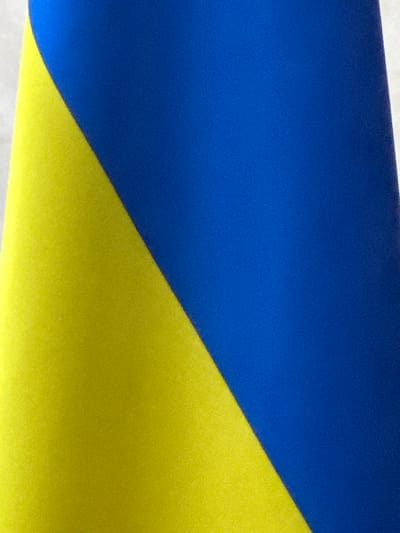 The media file is an abstract, close-up shot of two large swaths of fabric, one royal blue and the other bright yellow, meeting at a diagonal. The blue fabric occupies the upper left portion of the image, while the yellow fabric fills the lower right. The textured surfaces of both materials are visible, suggesting they might be some kind of textile or painted surface with a matte finish. There are no people, objects, or discernible scene depicted. The lighting appears even, with no strong shadows, suggesting an indoor setting or an overcast day. The colors are bold and vibrant, but there are no text elements present in the image. The abstract nature of the image, combined with the colors of blue and yellow, could be interpreted as reminiscent of the Ukrainian flag, especially given the provided location context of Myronivka, Ukraine.