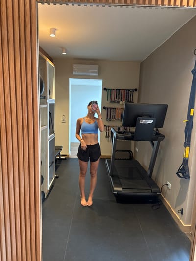 The scene depicts a female individual, presumed to be a young adult, taking a mirror selfie within a private gym setting located in San Luis Potosí City, Mexico. The subject stands barefoot in the center of the frame, clad in a light blue sports bra and black athletic shorts, holding a smartphone in her right hand to capture her reflection. Her face is partially obscured by the phone.
The environment features dark, square-tiled flooring and light brown-painted walls. Two white, cylindrical spotlights are affixed to the ceiling, and a white air conditioning unit is mounted on the upper wall. To the left of the subject, a wooden slatted partition borders the mirror, behind which a light-colored open shelving unit holds various gym equipment, including a visible black exercise ball. A black weight bench is also partially discernible. To the right, a black treadmill with a monitor displaying "MY TECHNOGYM" is positioned. Above the treadmill, multiple wall-mounted racks are densely populated with athletic medals. A yellow and black suspension trainer hangs from a wall anchor on the far right, with a small wall hook visible below it.