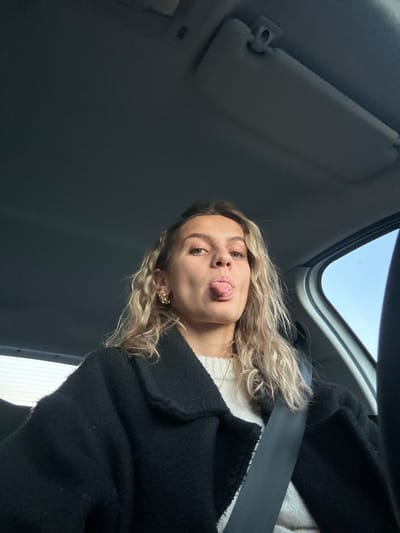 A woman with blonde, curly hair sticks her tongue out at the camera. She is wearing a black coat and a seatbelt is visible across her body. The photograph is taken from a low angle, showing the interior roof of a car. A sun visor is visible to the left of the image. The background shows a glimpse of sky through the car window. She has small gold earrings.