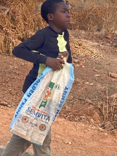 A boy dressed in a black shirt with yellow accents carries a large bag of Golden Penny Semvita Semolina. The bag is labeled "10 KG" and features a blue and white design. The boy is outside in a dry, arid landscape with brown soil and dried vegetation. He looks tired or contemplative as he holds the heavy bag. His clothing is casual, and he appears to be engaged in a chore.