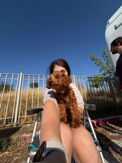 The image shows a young woman holding a small, brown, curly-haired dog while sitting in a portable chair. She is wearing a striped shirt. Her face is partially obscured by the dog. Behind her is a metal fence and dry, tall grass. A man in a dark shirt is visible in the background to the right, standing next to a white recreational vehicle. The scene is outdoors under a clear blue sky. The location is identified as Ribnik, Bulgaria.