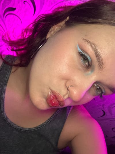 A young person with dark hair and fair skin, wearing a dark grey tank top, is pictured from the chest up. The person is leaning their head to the right, looking directly at the camera. Their lips are parted in a pout, highlighted with glossy red lipstick. They have light blue eyeshadow and eyeliner, with a small silver hoop in their nose. The background is a bright pink and purple patterned wall, likely illuminated by colored lighting. The image appears to be a selfie taken indoors, possibly in a bedroom. There is no indication of the location in Bishkek, Kyrgyzstan.
