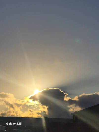 The image captures a dramatic sunset over Pątnów, Poland. The sun, a brilliant orb partially obscured by dark, heavy clouds, casts long, ethereal rays of light across the sky. These crepuscular rays are illuminated by the warm, golden hues of the setting sun, highlighting the edges of the clouds and creating a stark contrast with the darker, shadowed portions. In the foreground, the silhouette of rooftops suggests an urban or suburban environment. The lower left corner of the image contains the text "Galaxy S25," indicating the device used to capture the photograph. The sky transitions from a light, hazy blue at the top to a warmer, more intense yellow and orange near the horizon.