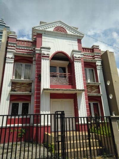A two-story building with classical architectural elements, painted predominantly red with white trim, columns, and decorative motifs, is depicted under a partly cloudy sky. The building is viewed from a low angle, looking upwards at its front facade.
The building's upper facade features a central white triangular pediment with a red semicircular detail, flanked by white balustrade-like parapet sections. The second floor includes a central recessed arched opening with a small balcony equipped with a black metal railing and white double doors behind it. This central section is framed by two large white fluted columns. To the left and right of the central balcony are rectangular windows with white frames and vertical mullions, each positioned above a band of light brown bricks and flanked by white pilasters.
The first floor presents a central entrance with white double doors, accessed via a small set of concrete steps. A flat white canopy extends over these doors. Similar to the second floor, this central section is flanked by white fluted pilasters. Rectangular windows identical to those on the second floor are present on the left and right sides of the first floor, also with white frames, vertical mullions, and sheer white curtains.
In the foreground, a black metal fence with vertical bars and a central gate separates the viewer from the building. Small green plants are visible near the base of the building and along the fence line. A paved or concrete area leads up to the building's steps behind the fence. An adjacent, taller bluish-grey building with a stepped roofline is partially visible to the left, and a tan-brown building with circular cutouts is partially visible to the right. No people, subjects, or animals are present in the scene. The sky above is blue with scattered white clouds.
The exact location within the city of Nathene, Laos, cannot be determined from the visual information provided in this image.