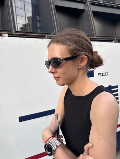 A young woman is depicted in a side-profile, half-length shot, leaning forward and looking downwards with focused attention. Her light brown hair is gathered in a casual messy bun, and she is wearing a black sleeveless top. Black, reflective sunglasses cover her eyes, partially obscuring her face, which shows a slight parting of her lips. A small, silver stud earring is visible in her left ear. On her right wrist, she wears a distinctive coiled silver-toned bracelet or watch with a red string detail. Her left hand gently holds a slender, silver-colored object, possibly a smartphone or other device, against her midsection.

The background on the right features a bright white surface marked with bold blue and red stripes, and the black letters "SCG" are prominently displayed. This suggests an outdoor sports area, such as a court or track. To the left, a modern building with dark, reflective glass windows is visible, mirroring reflections of other structures and the sky, indicating an urban environment. The overall lighting suggests it is daytime with clear weather conditions. The scene is set in Putuo District, China.
