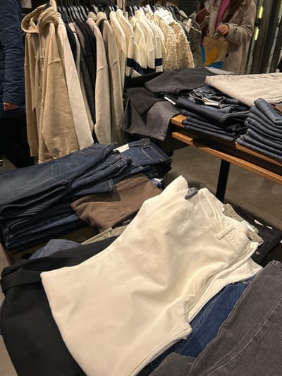 The image depicts an indoor scene within a clothing store, likely located in Chalkida, Greece, showcasing a variety of folded garments. The shot is taken from a slightly elevated, tilted perspective, looking down onto display tables and wooden shelves.
The primary subjects are numerous articles of clothing. In the foreground and mid-ground, there are multiple piles of folded trousers, predominantly jeans, in various washes and colors including dark blue, black, white, grey, and brown. A large stack of pristine white or off-white trousers is prominently featured on the right side of the frame. Towards the left, a tall stack of folded tops, sweaters, and shirts is visible, mainly in neutral tones such as cream, white, beige, and light grey. Some striped garments, including a cream and dark blue striped top, are present within this stack. A piece of cream-colored lace fabric is also seen on top of one of the piles.
In the blurred background, two individuals are partially visible. One person on the far left appears to be wearing a light-colored coat, possibly engaged in browsing. Another person with dark hair is also indistinctly seen. Their faces and emotions are not discernible.
The scene suggests an activity of shopping or browsing in a retail environment. The clothes are arranged on wooden shelves and flat surfaces, typical of store displays. Small white tags or labels are attached to some garments, but the text on them is not legible. The lighting is artificial, indicative of an indoor setting, and there are no specific cues regarding the time of day or weather.