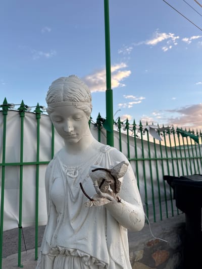 The image shows a white statue of a woman in classical attire, positioned outdoors. The statue appears weathered, with peeling paint and visible cracks. Her left hand is extended, holding a broken piece of what seems to be rebar and concrete, suggesting damage. Behind the statue is a green metal fence with pointed finials, topped with decorative lights. Beyond the fence, a white fabric or tarp is visible, and further back, a black trash bin. The sky above is a clear blue with scattered clouds tinged with sunset colors. The scene is set in Sunland Park, United States of America.