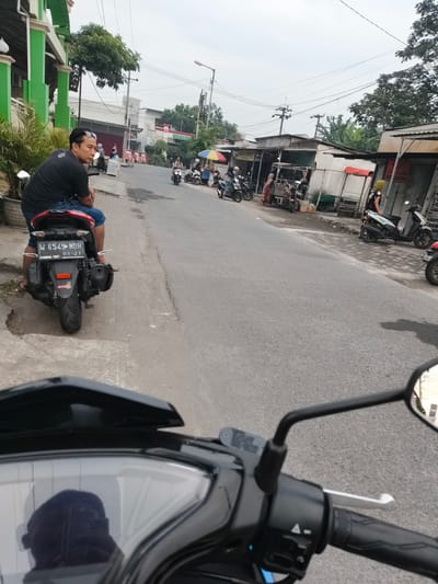A man wearing a black t-shirt and blue shorts is sitting on a red motorcycle parked on the side of a street in Sidorejo, Indonesia. He is looking to the left, and his sunglasses are on top of his head. The motorcycle has a license plate that reads "W 6549 NDH". In the background, there are several shops and other motorcycles parked along the street. The sky is overcast, and the road is wet, suggesting recent rain. The scene appears to be a typical street view in a residential or commercial area.