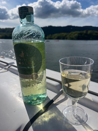 The image depicts a bottle of Wise Wolf Sauvignon Blanc 2023, lying horizontally next to a stemmed plastic glass partially filled with the same white wine. The scene is set outdoors, likely on a boat or dock, as the background reveals a body of water, treed mountains, and a sky with scattered clouds. The bottle is mostly full and has condensation on its surface. The glass is placed to the right of the bottle, casting a shadow. The surface on which the bottle and glass rest is white. The bottle's label is visible, indicating the brand, type of wine, and origin (Pays d'Oc, France). The label also states that the bottle is made using 100% post-consumer recycled glass. There are no people visible in the image. Given the background, the image could possibly be located in Bowness-on-Windermere in the UK.