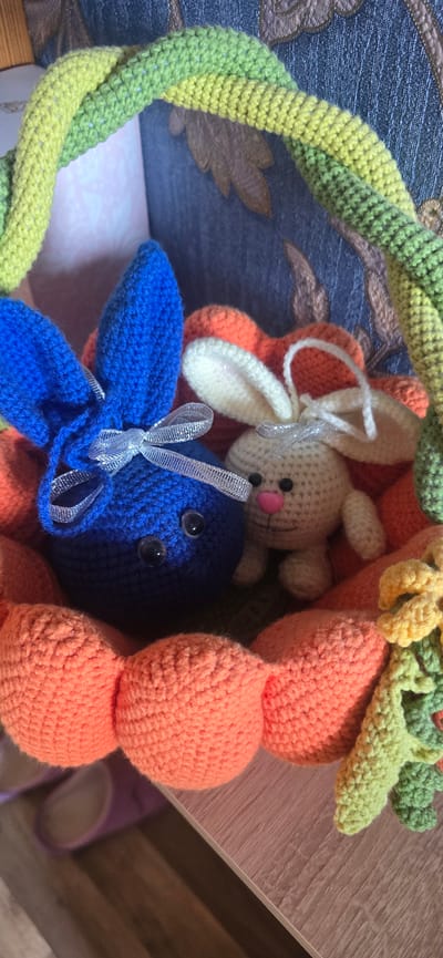 This image displays a close-up view of a collection of crocheted Easter-themed decorations. The main subjects are two crocheted bunnies, one predominantly blue and the other a lighter white color. They are nestled amongst other crocheted elements, including what appear to be large orange petal-like shapes and green and yellow braided loops, possibly forming a wreath or garland.
The blue bunny is positioned in the foreground, with its ears prominent and adorned with a silver ribbon bow. It has googly eyes. The white bunny is partially visible behind the blue one, also featuring googly eyes and a small pink nose.
The crocheted textures are highly detailed, showcasing the stitchwork. The color palette is bright and cheerful, dominated by blues, oranges, yellows, and greens, suggestive of spring and Easter. The items are placed on what appears to be a wooden surface, possibly a shelf or table, with a textured blue fabric background visible behind the decorations. The lighting suggests an indoor setting, possibly during the daytime. There is no visible text in the image. The overall mood conveyed is one of handmade crafts and festive celebration.
