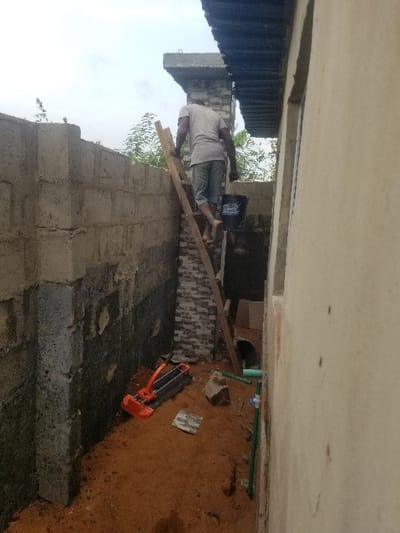 The image displays an outdoor construction or renovation site in Edu Town, Nigeria, situated within a narrow passage between two structures. A male individual, observed from behind, wearing a light-colored t-shirt and dark shorts, is barefoot and ascending a crude wooden ladder. He carries a dark bucket in his left hand. The ladder rests against a partial wall clad with grey and white patterned bricks. Above this structure, a concrete ledge and a dark corrugated roof overhang are visible against an overcast sky. To the immediate left, an unfinished concrete block wall, darkened and stained at its base, borders the passage. To the right, a light-colored, likely plastered, wall forms the opposing boundary. The ground consists of bare, reddish-brown soil. An orange and black manual tile cutter, several rocks, fragments of material, and green and white PVC pipes are scattered on the soil. Sparse green vegetation is visible above the left wall and in the distant background.