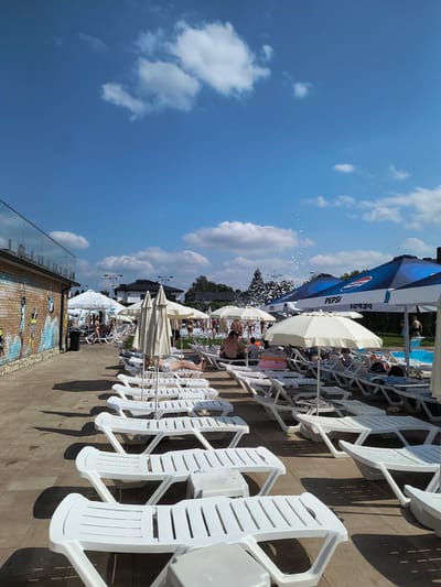 This image depicts an outdoor scene at a water park in Velyka Omeliana, Ukraine, on a clear, sunny day with scattered white clouds. The foreground is dominated by rows of empty white plastic lounge chairs arranged on a paved surface made of square tiles.
In the middle ground, several large, open umbrellas provide shade over some of the lounge chairs, some of which are occupied by people. A prominent blue umbrella with a white Pepsi logo is visible. Further back, a group of people are gathered, some of whom appear to be in or near a pool, with foam or bubbles rising into the air. A diving board is also visible near the pool.
To the left, a low brick building with a glass railing on top is seen. The brick wall features colorful graffiti depicting stylized figures. Trees are visible in the background, along with buildings and lampposts. The overall impression is one of a recreational area during a daytime event or normal operating hours.