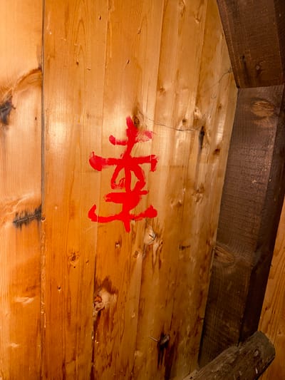 The image shows a section of a wooden structure, likely an interior wall or ceiling, composed of light-colored, horizontal planks. A symbol or word is painted in red on the planks, appearing as four distinct strokes with varying widths. The wood is aged, with visible knots and imperfections. The location is not specified as Rila, Bulgaria in the provided image.