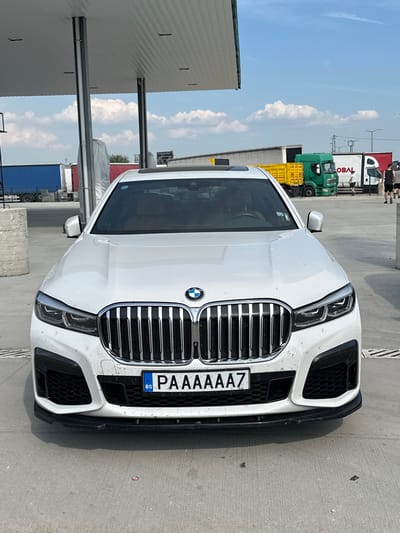 The scene is located in Edirne Merkez, Turkey. A white BMW G70 7 Series sedan is positioned facing the camera under a white corrugated metal gas station canopy. The vehicle exhibits a large, chrome vertical kidney grille flanked by LED headlights and black lower bumper grilles with a black front splitter. The front bumper and lower bodywork are lightly soiled with dirt or dust. The front license plate, centered on the bumper, displays "BG PAAAAAZ", indicating Bulgarian registration. To the immediate left of the car, a cylindrical grey concrete bollard is present.

The ground is composed of grey concrete pavement, with a drainage grate visible near the car's front right wheel. The overhead canopy structure is supported by dark grey vertical columns.

In the background, under a bright blue sky with scattered white cumulus clouds, a truck parking area or industrial lot is visible. Multiple commercial vehicles are present, including a blue semi-trailer on the far left, followed by a grey semi-trailer. Further to the right, a green cab-over truck with a yellow flatbed trailer is parked. Adjacent to it is a white semi-trailer truck featuring a partially visible red "GLOBAL" logo on its side.

Three males are observed in the mid-ground near the parked trucks. One male, wearing a white t-shirt and dark shorts, is walking rightwards between the green and white trucks. Two additional males, both wearing dark t-shirts and shorts, are walking rightwards further behind the white truck; one faces away from the camera, while the other faces towards it.