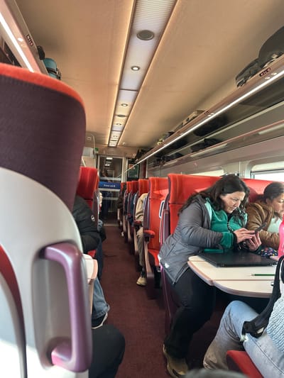 The image displays the interior of a passenger train carriage, viewed from a slightly elevated position looking forward down the central aisle. The carriage is equipped with multiple rows of high-backed seats upholstered in red fabric with purple-grey accents, arranged in a 2+1 configuration. A reddish-purple carpet covers the aisle floor. Overhead luggage racks, featuring integrated linear LED lighting, line both sides of the carriage; several pieces of luggage, including a light blue hardshell suitcase, are stowed within them. The ceiling is light-colored, fitted with recessed circular lights and a central illuminated perforated panel. Natural light enters from windows on the right side of the carriage.
Approximately seven to ten individuals are visible within the carriage. In the mid-ground on the right, a woman with dark hair, wearing a grey quilted jacket over a teal garment, is seated at a shared table. She is actively using a dark-colored smartphone held in both hands, looking down at its screen. A closed black laptop and a green pencil are positioned on the table in front of her. To her right, another woman is partially visible, also appearing to look down. Other passengers are seated along the aisle, primarily facing forward, including one person in a black puffer jacket on the mid-ground left. At the far end of the visible carriage section, a glass door with a "Free wi-fi" decal separates it from an adjacent area.
There are no discernible visual elements within this image that definitively establish the train's location in the city of Breda, Netherlands.