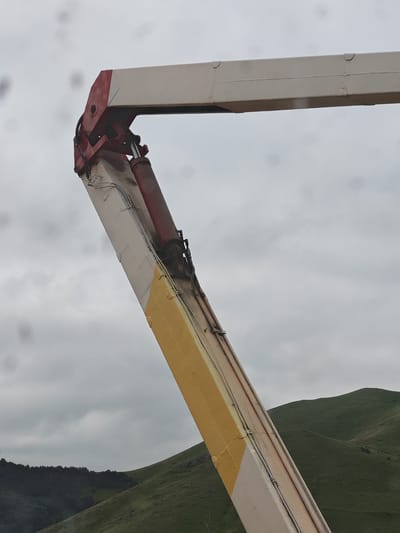 A close-up, eye-level, outdoor shot captures a section of a crane's mechanical arm against a cloudy sky. The arm is painted in alternating sections of white, red, and yellow, with visible bolts and hydraulic components. The scene is situated in Bazum, Armenia, with rolling green hills and a dense forest visible in the background. The overall mood is overcast and industrial.