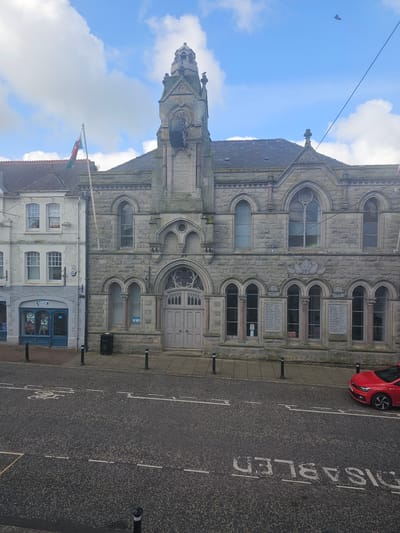 The image captures a street scene in Holywell, United Kingdom. In the foreground, there is a road marked with "DISABLED" text, indicating a parking spot for people with disabilities. A red car is parked on the side of the road.
Adjacent to the road, a building with a stone facade dominates the scene. The building's architecture is notable for its Gothic Revival style, featuring arched windows, intricate stonework, and a tall tower crowned with a spire. Plaques bearing names of historical significance are attached to the building, possibly serving as a war memorial or a "Roll of Honour".
Next to the substantial stone building, a smaller, white building with a blue storefront is visible. A Welsh flag is flying atop this smaller building. The sky above is partly cloudy with blue sky visible.