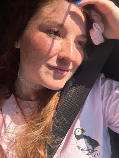 A close-up, high-angle outdoor shot shows a young woman with auburn hair and freckles wearing a pink t-shirt in what appears to be a vehicle. She is wearing a white choker necklace and a pink scrunchie is attached to her hair. The sun is shining through the window, casting shadows on her face. A seatbelt is visible across her chest. The t-shirt has a graphic of a puffin and text that appears to be in Cyrillic script. The location is identified as the city of Крутобереговый, Russia.