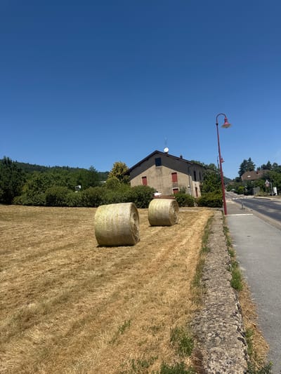 The image shows a rural scene in Coteaux du Lizon, France, on a clear day. In the foreground, a field of dry, golden hay is visible, with two large, round hay bales resting on it. The hay field is bordered by a low stone wall on the right, beyond which is an asphalt road with a sidewalk. A red lamppost with a single light fixture stands beside the road. Further back, a light-colored, two-story house with red shutters is situated among trees and bushes. A line of green trees forms a backdrop behind the house, with a few more houses visible in the distance along the road. The sky is a vibrant, cloudless blue.