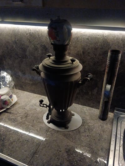 A vintage samovar, an object used for heating and boiling water, sits on a speckled countertop in what appears to be a dimly lit kitchen or dining area. The samovar is dark in color and has ornate handles. To the left of the samovar, a small plate holds two teacups, one of which is partially visible and decorated with floral patterns. Next to the teacups is a crumpled piece of aluminum foil. To the right of the samovar, a tall, metallic cylinder, possibly a pipe or a part of a chimney, stands upright. The countertop is wet in several places, with water droplets reflecting the overhead lighting, which appears to be an LED strip running along the wall behind the samovar. The wall itself is textured and dark. In the foreground, a portion of a stainless steel sink with visible water is present. The overall scene suggests a domestic setting, potentially in Türkan, Azerbaijan, given the context provided, where such samovars are traditionally used.