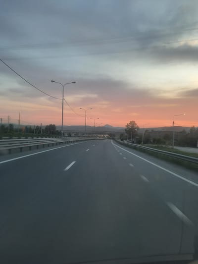 This media file depicts a roadside scene at dusk, as suggested by the muted light and the gradient of orange and pink in the sky. The perspective is from a moving vehicle on a highway, indicated by the blurred asphalt road and the dynamic lines of the road markings. The highway runs parallel to a metal barrier and a line of streetlights. On the left side of the road, a vast sky dominates, filled with soft, diffused clouds. In the distance, silhouetted mountains form the horizon, partially obscured by trees and buildings, suggesting a semi-rural or suburban setting. The overall mood is tranquil and expansive, capturing the fleeting beauty of twilight. The time of day is clearly dusk, with the sky displaying vibrant hues of orange and pink. There is no visible text within the image.